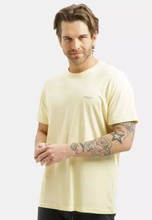 Mężczyzna z wystylizowanymi brązowymi włosami, ubrany w jasnżółtą koszulkę, pokazujący wytatuowane prawe przedramię z różami i gwiazdami na jednolitym tle.