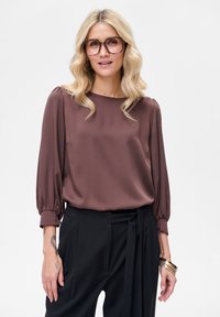 Brązowa bluzka z długim rękawem z gładkiej tkaniny, z marszczonymi mankietami i luźnym krojem, połączona z czarnymi spodniami z wysokim stanem.