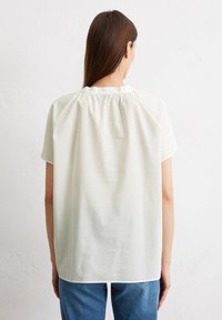 Helles beige Kurzarm-Blusenshirt mit gerafftem Rückenkragen und lockerer Passform, kombiniert mit blauen Jeans. Glattes, leichtes Material.