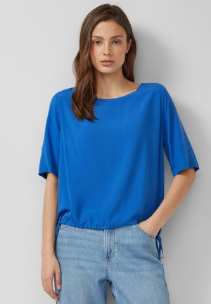 Kobieta o brązowych włosach, ubrana w luźny krótkim rękawem top w kolorze królewskiego niebieskiego i jasnoniebieskie dżinsy, stojąca na jednolitym jasnoszarym tle.