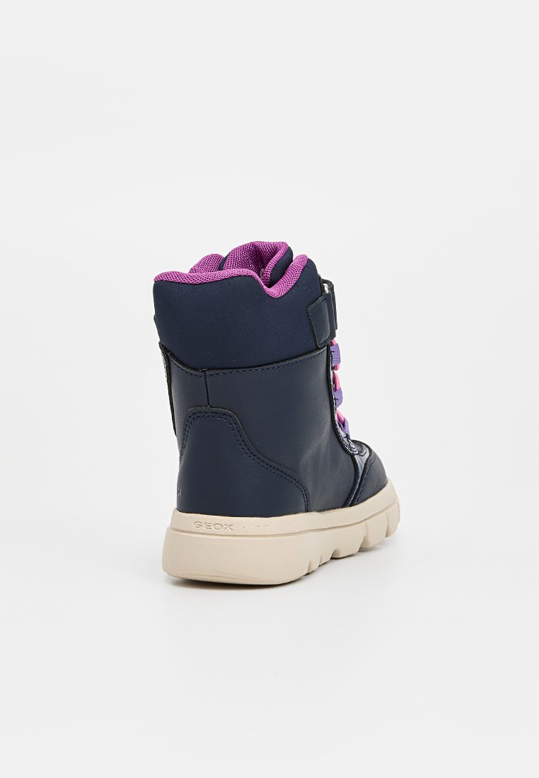 Dunkelblaue High-Top-Sneaker mit lila Innenfutter, pinken Schnürsenkeln, beiger Sohle und Riemen an der Seite, schräg dargestellt, um die Rückseite und die Außenseite zu zeigen.