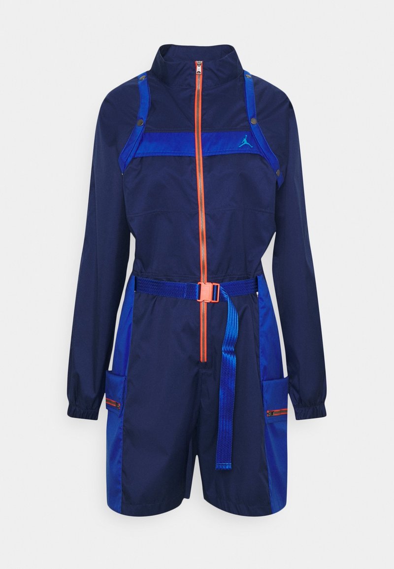 Combinaison bleu marine à manches longues, col montant, fermetures éclair orange et accents bleus. Comprend une ceinture ajustable et plusieurs poches.