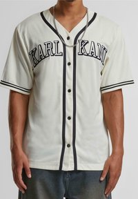 Biała koszula na krótki rękaw z guzikami, z czarnymi akcentami, z napisem "KARL KANI", teksturowaną tkaniną oraz czarnymi paskami na kołnierzu i krawędziach rękawów.