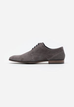 Chaussure oxford en daim gris avec un bout pointu, des détails cousus et un talon en bois, présentant un design classique et minimaliste.