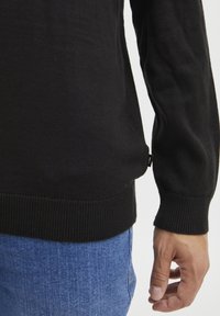 Zwarte gebreide trui met geribde manchetten en zoom, met een label aan de zijkant. Draagt over blauwe denim jeans.