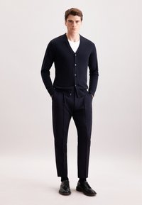 Cardigan blu navy con scollatura a V, chiusura a bottoni e maniche lunghe abbinato a pantaloni scuri e scarpe eleganti nere su uno sfondo neutro.