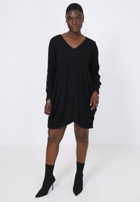 Zwarte oversized jurk met een V-hals, lange mouwen met gouden knopen, en een comfortabele pasvorm, gestyled met zwarte enkellaarsjes.