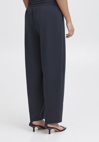 Pantalon large bleu marine avec une ceinture élastique, une texture lisse, des poches latérales et une coupe ample, associé à des sandales à talons noires.