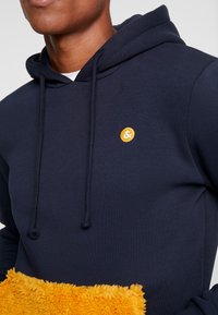 Man som bär en marinblå huvtröja med dragsnören och en liten gul cirkulär logotyp på bröstet, håller i ett lurvigt gult föremål.