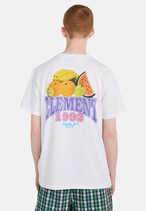 Persona con capelli rossi corti indossa una maglietta bianca con una grafica colorata di frutta e il testo "ELEMENT 1992 Atlanta GA USA" sulla schiena.