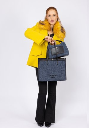 Cappotto giallo con colletto di pelliccia, pantaloni neri a zampa, tiene una grande borsa a tracolla blu texture e una piccola borsa a mano con dettagli neri.