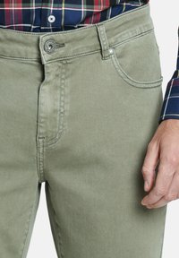 Pantalones de algodón verde oliva con una textura suave, con un bolsillo frontal, costuras en contraste y un cierre de botón metálico.