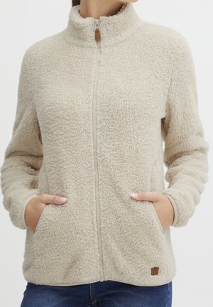 Kvinde iført en beige fleecejakke med lynlås og hænder i forlommerne, kombineret med blå jeans, på hvid baggrund.