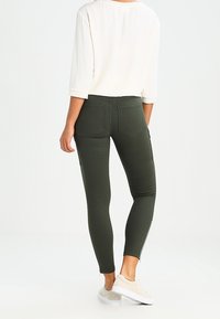 Pantalones ajustados verde oliva con cremalleras en los tobillos, que cuentan con dos bolsillos traseros y una textura suave, combinados con una blusa blanca.