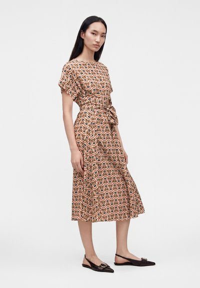 Femme debout portant une robe midi à motifs avec ceinture, manches courtes et des ballerines noires pointues à bride arrière, sur un fond uni.