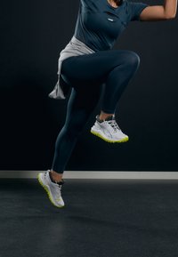 Bílé a šedé sportovní boty s neonově žlutými akcenty, spárované s tmavě modrými legínami a šedým svetrem uvázaným v pase.