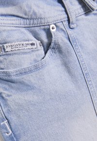 Τσέπη από ανοιχτό μπλε τζιν με ένα μικρό ετικέτα που γράφει "Juicy Couture Jeans", με ασημένιους αμπράδες και εμφανείς ραφές.