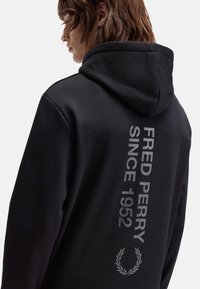 Sudadera con capucha negra que presenta un destacado gráfico de texto gris con la inscripción "FRED PERRY SINCE 1952" y un diseño de laurel en la parte posterior.