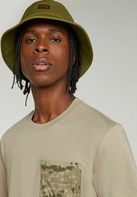 Beige T-shirt met een gestructureerde camouflagezak. Olijfgroene bucket hat met een klein logo-patch, gedragen door een persoon met gevlochten haar.