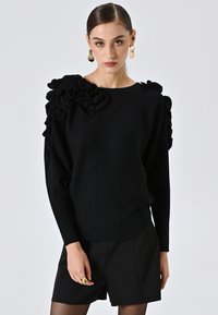 Maglione nero con dettagli a volant sulle spalle, scollatura rotonda e maniche lunghe. Indossato con pantaloni corto neri sartoriali e calze trasparenti.