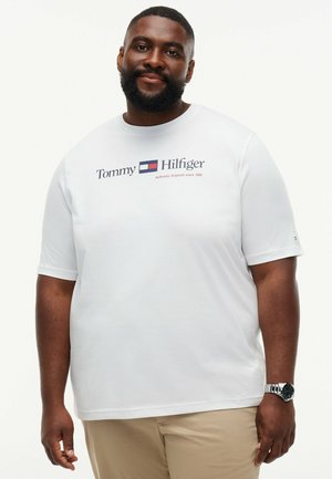 Mand med skæg iført en hvid Tommy Hilfiger t-shirt og beige bukser, stående med et neutralt udtryk mod en ensfarvet baggrund.