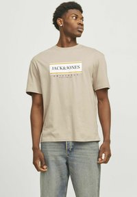 Beige Baumwoll-T-Shirt mit Rundhalsausschnitt und kurzen Ärmeln, das auf der Vorderseite ein grafisches Logo-Design in Schwarz und Gelb aufweist.