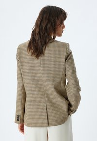 Blazer su misura in una fantasia a quadri marrone e beige, con uno spacco singolo, spalle strutturate e bottoni a contrasto sui polsini.
