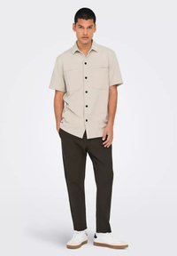 Camisa corta de color beige claro con dos bolsillos frontales y botones negros, combinada con pantalones oscuros y zapatillas blancas. Textura suave.