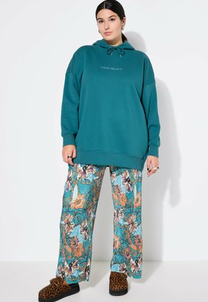 Kvinde med mørkt hår iført en stor, turkis hættetrøje, vide bukser med blomsterprint i blå og orange nuancer og leopardmønstrede tøfler, der står mod en hvid væg.