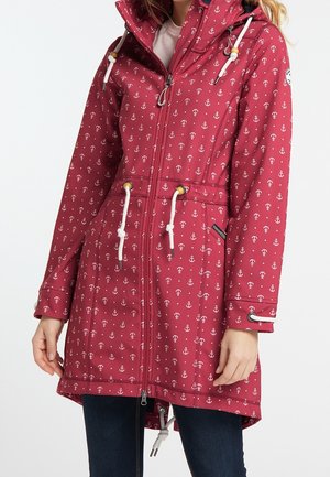 Femme portant un imperméable à capuche rouge orné de motifs d'ancres blanches, avec des cordons blancs et un jean bleu foncé.