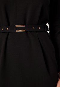 Czarna sukienka z dopasowaną górą, plisowanymi detalami oraz paskiem w talii z metalowymi akcentami i dwiema prostokątnymi złotymi elementami.