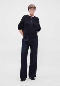 Ναυτικό πλισέ πλεκτό μπουφάν με κουμπιά μπροστά, ριγωτή υφή και χαμηλούς ώμους, συνδυασμένο με ριγέ παντελόνι σε φαρδιά γραμμή και μαύρες γόβες.