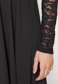 Vestido preto com um corpo de tecido suave e mangas longas de renda com padrões florais. Mão segurando o tecido perto da costura.