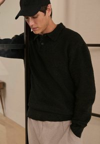 Sort fleece pullover med krave, der har to knapper foran, struktureret stof og relaxed fit, parret med lysegrå bukser.