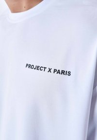Witte katoenen t-shirt met de tekst "PROJECT X PARIS" in zwarte borduursel, korte mouwen en een ronde hals. Glad textuur.