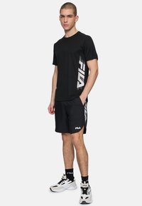 Camiseta deportiva negra con el logo blanco de "FILA", combinada con pantalones cortos negros con laterales abiertos y acentos de logo en blanco. Zapatillas blancas completan el look.