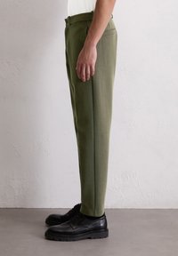 Pantalons ajustés vert olive avec une coupe droite, poches latérales et texture lisse, accompagnés de chaussures noires à lacets.