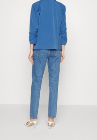 Modrá bunda se stahovanými rukávy nošená přes světle modré džíny. K outfitu patří bílé sandály na podpatku. Neutrální pozadí.