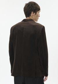 Bruine suède blazer met een getailleerde pasvorm, notch revers en een enkele rugopening. Soepele textuur met knoopmanchetten.