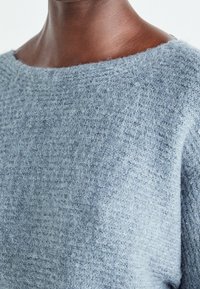 Světle modrý svetr s texturovaným pleteným vzorem, širokým výstřihem a měkkým, huňatým materiálem, který částečně pokrývá rameno.