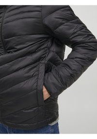 Persona que lleva una chaqueta acolchada negra con un bolsillo lateral, con la mano metida, combinada con jeans azules y una camiseta gris debajo.