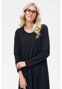 Czarna sukienka z długim rękawem, okrągłym dekoltem, rozszerzanym dołem i luźnym krojem. Modelka nosi okulary i kolczyki z jasnoniebieskimi akcentami.