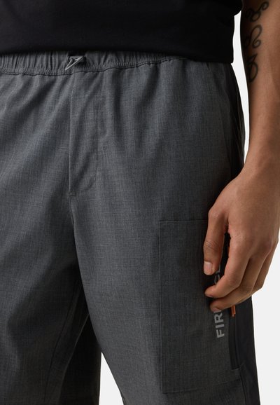 Zbliżenie osoby noszącej ciemnoszare spodnie z elastycznym paskiem w talii i boczną kieszenią z suwakiem oraz częściowym białym napisem.