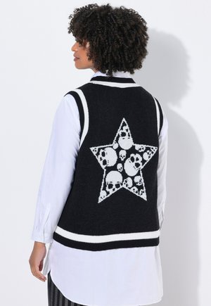 Une personne aux cheveux bouclés porte un gilet sans manches noir orné d'une étoile blanche remplie de motifs de skulls, par-dessus une chemise à manches longues blanche.