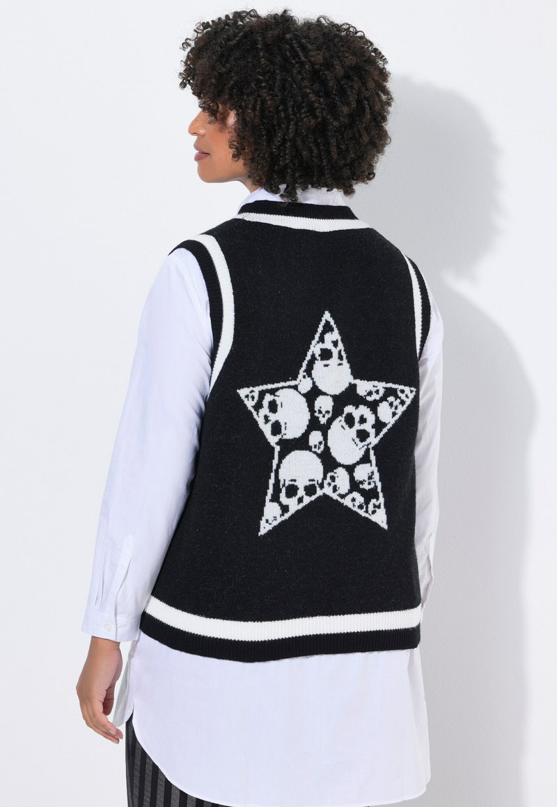Persona con capelli ricci indossa un gilet senza maniche nero con una stella bianca piena di scheletri, sopra una camicia a maniche lunghe bianca.