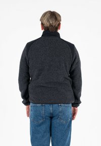 Zwarte fleece jas met een gestructureerd, fluffy oppervlak en zwarte schouderaccenten, voorzien van een hoge kraag en elastische manchetten.