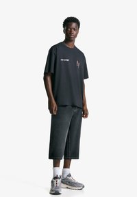 Svart bomullströja med "TOY STORY" text och grafik, kombinerad med mörka denimkortbyxor och grå sneakers med vita strumpor.