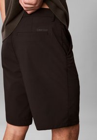 Shorts marron en tissu lisse, avec une poche arrière et un logo brodé sur la ceinture. Coupe ajustée avec un ourlet net.