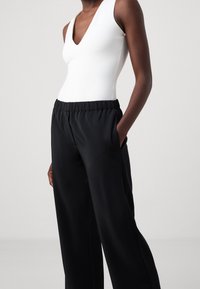 Vit ärmlös bodysuit med djup V-ringning, i kombination med svarta byxor med vida ben som har resår i midjan och sidofickor.