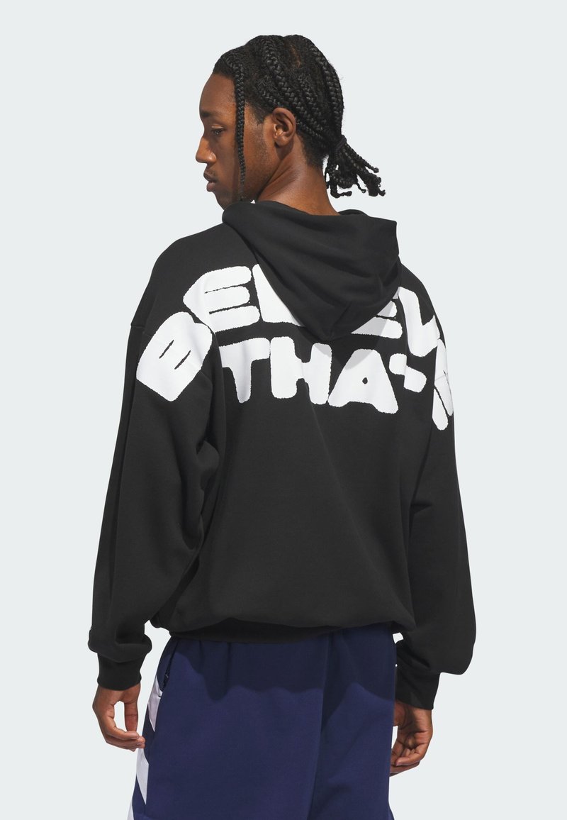 Άνδρας με πλεκτά μαλλιά που φοράει μαύρο φούτερ με μεγάλο λευκό κείμενο στην πλάτη, συνδυασμένο με σκούρο μπλε αθλητικό παντελόνι με λευκές ρίγες.